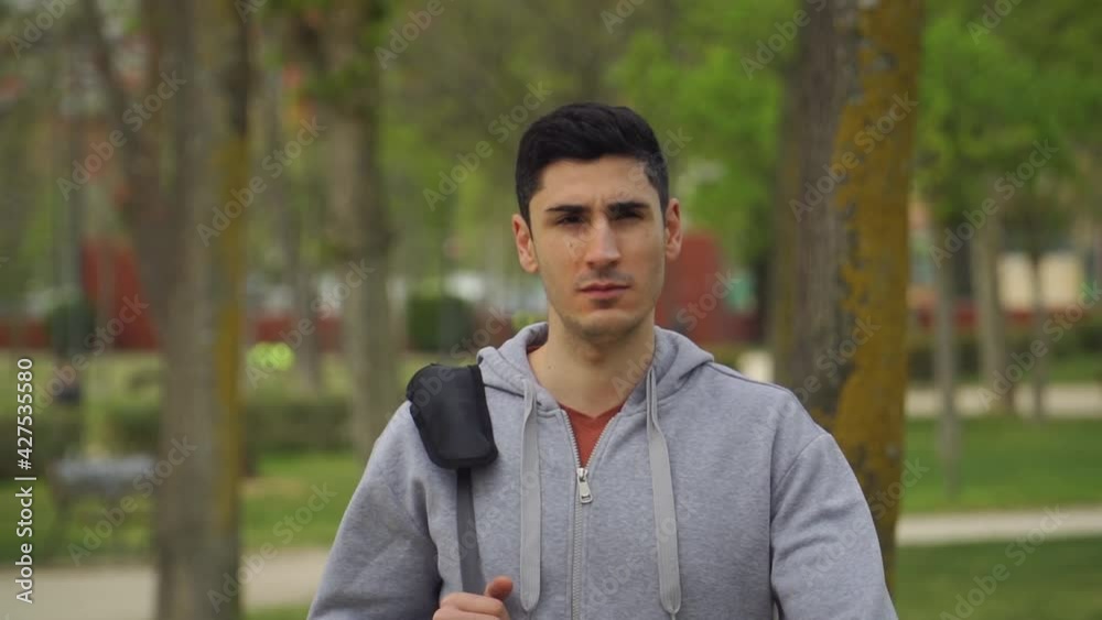 Hombre de pelo negro caminando a cámara lenta con su bolsa de deporte, hombre con sudadera gris y chándal caminando, deportista caminando por el parque, caminando de  frente a la cámara 