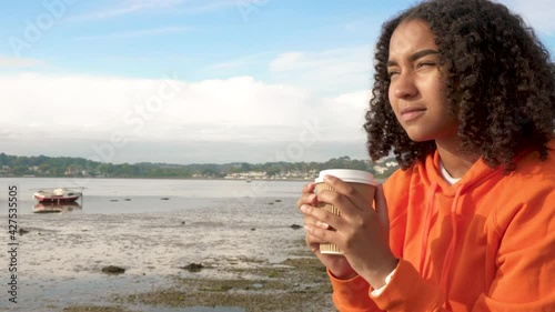 Beautiful mixed race African American girl teenager young woman wearing orange hoodie, drinking takeout coffee by the sea looking sad or thoughtful then happy smiling