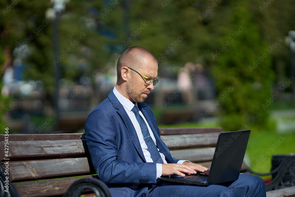 The man uses a laptop to work outdoors. He is sitting on a bench. green foliage in the background The man uses a laptop to work outdoors. He is sitting on a bench. green foliage in the background