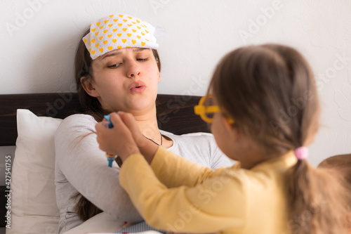 Cute little girl wearing uniform playing doctor or nurse with young mum or nanny in bedroom, makes an injection, measures temperature, family spending leisure time at home together, role play game