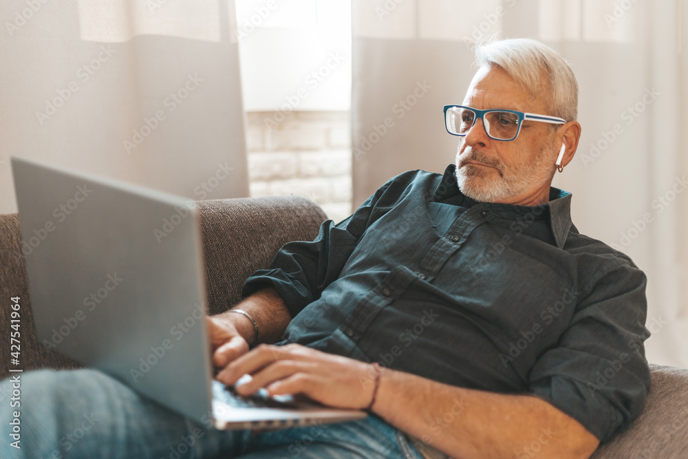 Focused on work at home. An adult man is watching a video on a laptop and is lying on the couch. Remote work, a pensioner uses a computer. Focused on work at home. An adult man is watching a video on a laptop and is lying on the couch. Remote work, a pensioner uses a computer.