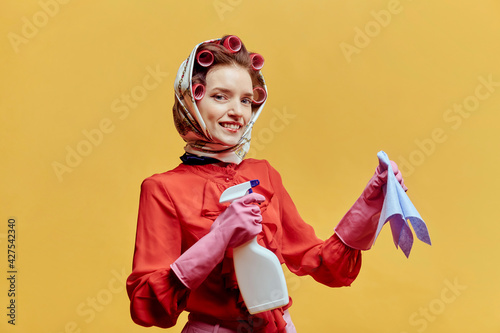 Happy cleaning lady wearing hair curlers and a headscarf. In the hands of a spray for washing windows and a rag. He looks at the camera. Yellow background. Pin-up style.