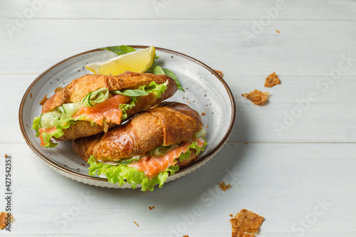 Two croissant sandwiches with lightly salted salmon and lettuce leaves, on a gray plate. Close-up, light wooden background with crumbs