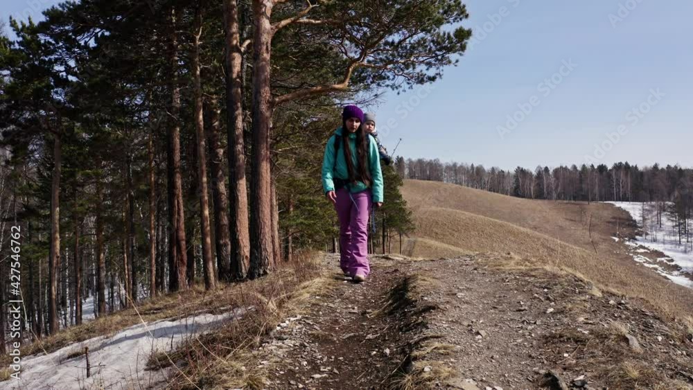 A young mother carries her child in a backpack along a forest trail in the mountains in spring A young mother carries her child in a backpack along a forest trail in the mountains in spring