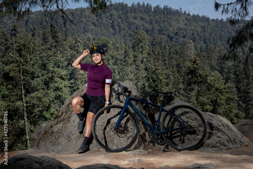 Chica ciclista recargado de una piedra con su bicicleta al lado, con un paisaje al fondo. Deporte al aire libre