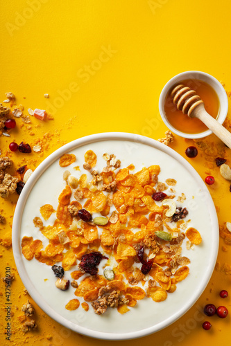 Breakfast concept. Cornflakes, nuts, seeds, berries and honey in a bowl with milk on a bright yellow background. Flakes and nuts scattered on the background. Healthy food. Top view. 