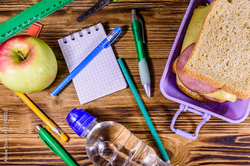 Bottle of water, ripe apple, different stationeries and lunch box with sandwiches on a wooden table. Top view