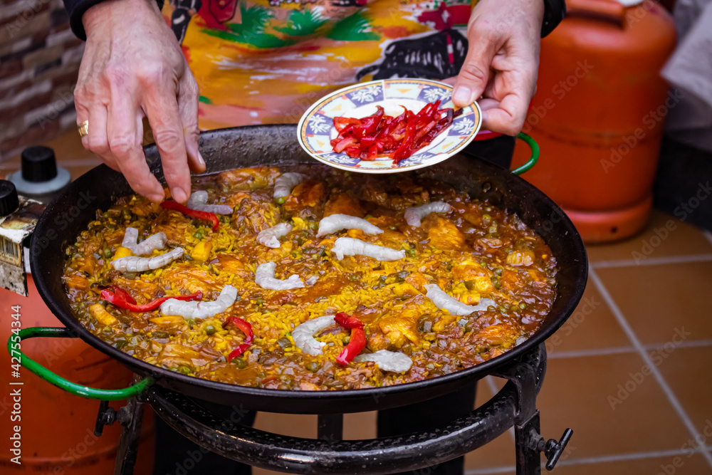 Fototapeta premium Chef hands cooking a paella in a paella pan. Chef putting red pepper on the yellow rice. Cooking Spanish traditional food in a butane gas pan. Andalusian paella recipe.