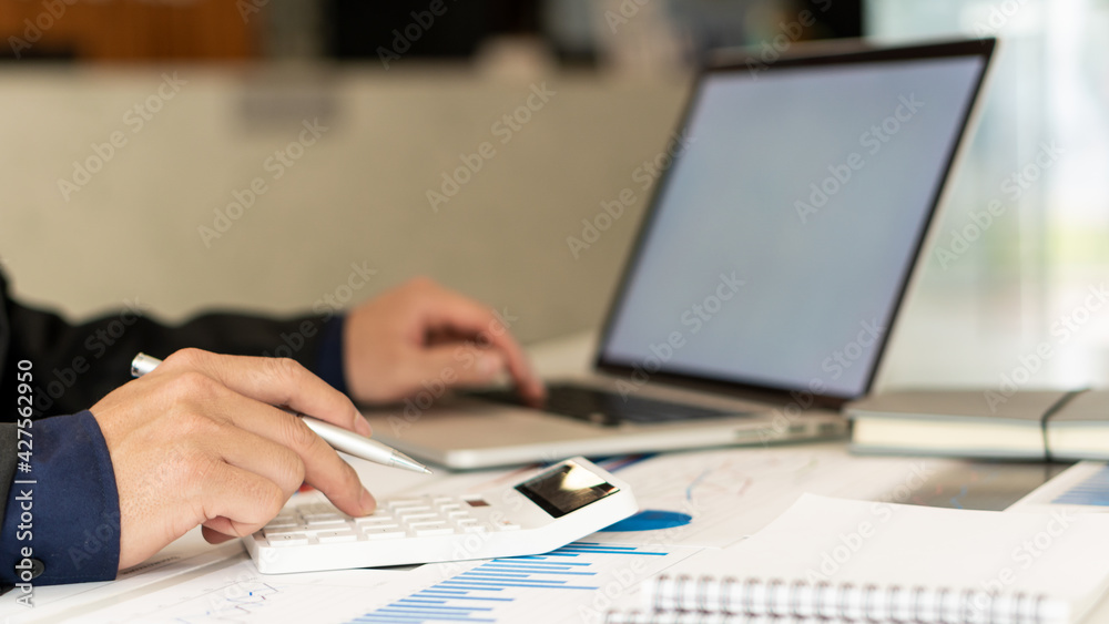 A businessman working on a desk with documents, graphs, charts, calculators and a laptop computer.