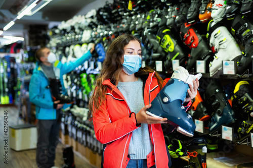 Positive female buyer in a medical protective face mask chooses ski boots in a sports equipment store