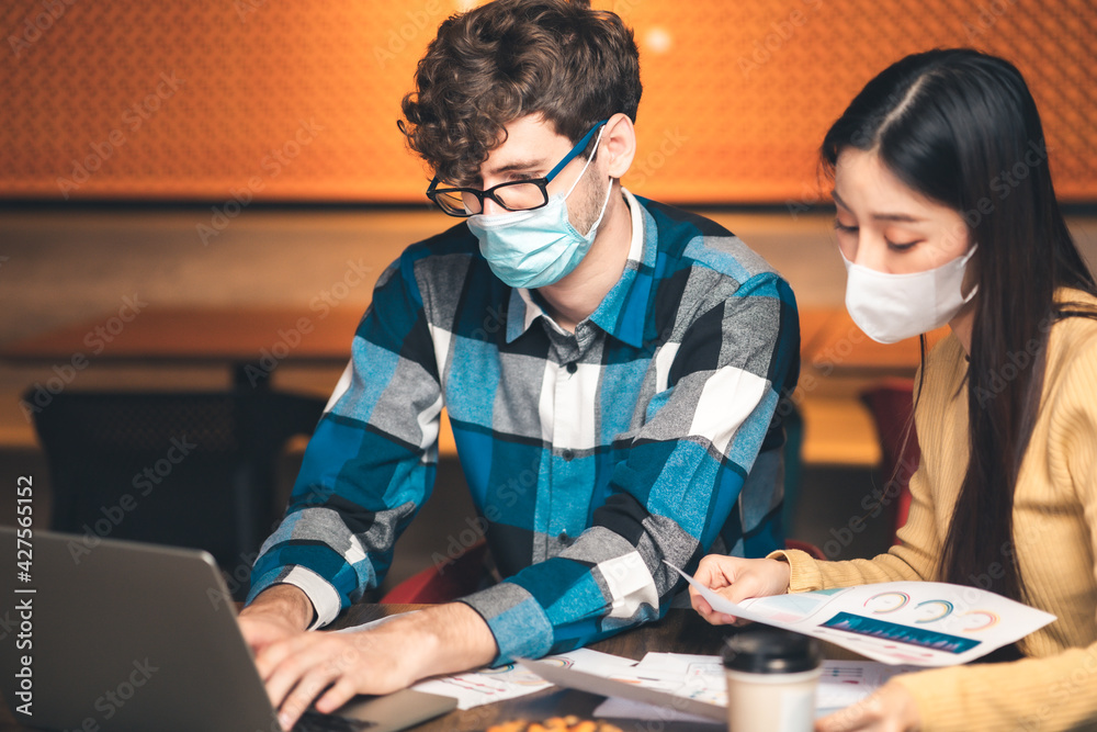 Naklejka premium young asian woman and caucasian man Business people Working discuss finding new strategy solution.People putting protective face mask discussion business plan using laptop at cafe.