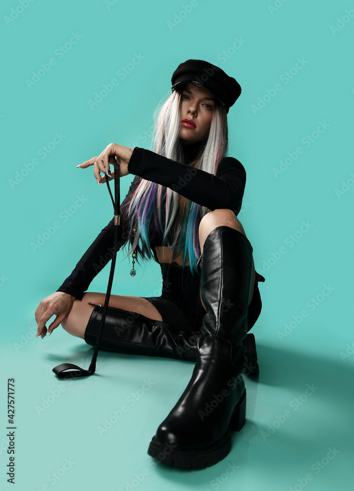 Foto Stock Young blonde bdsm woman mistress with colored strands in black  leather sexy costume and high boots sits on floor with horse whip, riding  crop. Sexual games, bdsm, glamour concept