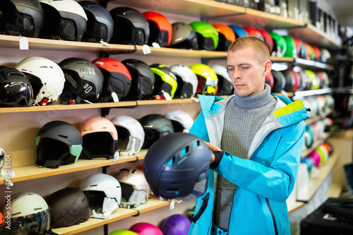 American man buyer in blue ski jacket chooses helmet in a sports equipment store