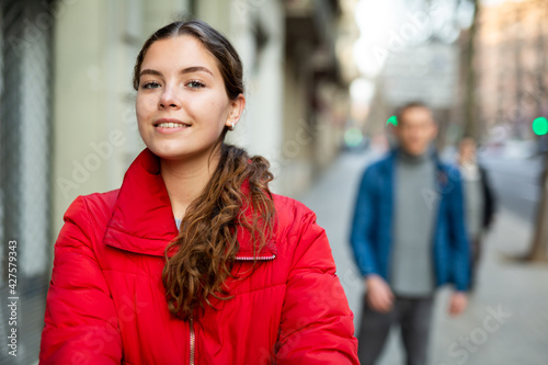 Portrait of young positive smiling woman in a red jacket walking on the street of the city