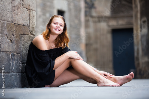 Canvas Print .Positive young girl sitting barefoot near the stone wall at the street
