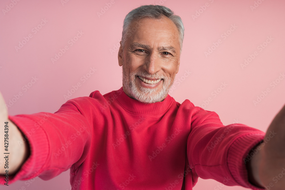 Close-up shot of a senior man's hands holding his phone, taking a selfie, on a pink background. Smiling man isolated on pink background. Close-up shot of a senior man's hands holding his phone, taking a selfie, on a pink background. Smiling man isolated on pink background.