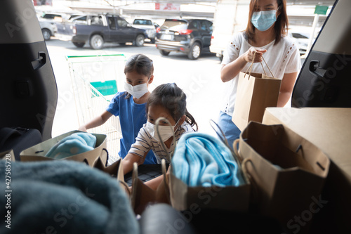 Asian mother and two daughters wearing protection mask helping to put stuffs in the shopping trolley into trunk of car together in parking during coronavirus pandemic as new normal lifestyle.
