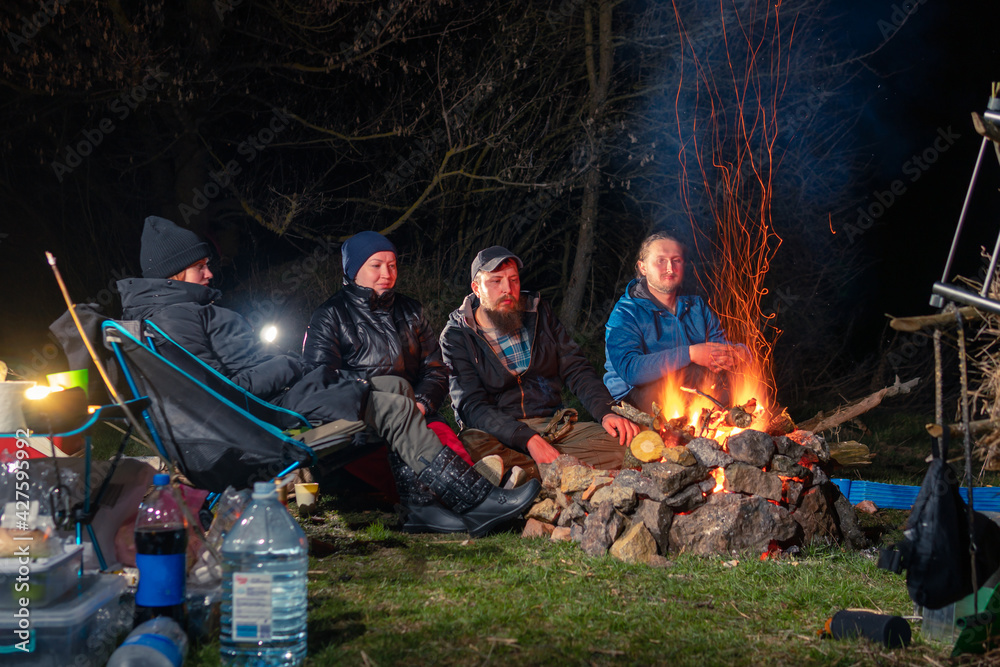 Group of tourists relaxing near campfire after a trip, Lighting tents, camping equipmrnt. Night spring time. Front view Group of tourists relaxing near campfire after a trip, Lighting tents, camping equipmrnt. Night spring time. Front view