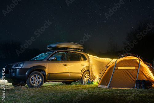 Night landscape with illuminated tent and 4wd car, lights and stars in the background. Spring camping. Tourism and vacation. Roof rack, car luggage box