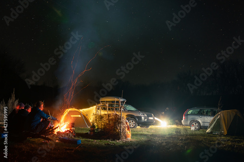 Group of travelers sitting near bonfire  cars and camp tent under beautiful night sky. View of blue starry sky over meadow with travelers near campfire.