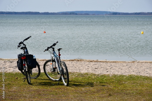 Close up on two bikes with bags standing next to a sandy coast of a river or lake with some red and yellow buoys visible in the distance seen on a sunny spring day on a Polish countryside 