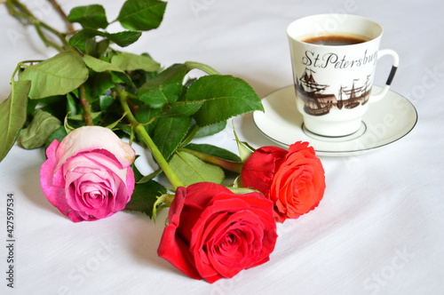 A white cup with black coffee is on a saucer. Nearby lies a bouquet of three beautiful multi-colored roses with green leaves. On a white linen tablecloth.