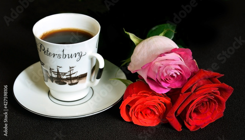 A white cup with black coffee is on a saucer. Nearby lies a bouquet of three beautiful multi-colored rose buds. On a dark brown background.