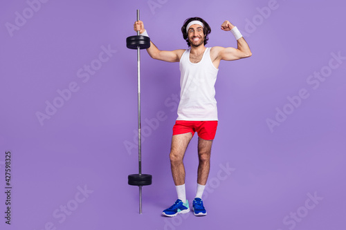 Full size photo of young funky smiling man in glasses brunette hair showing muscles hold barbell isolated on purple color background photo