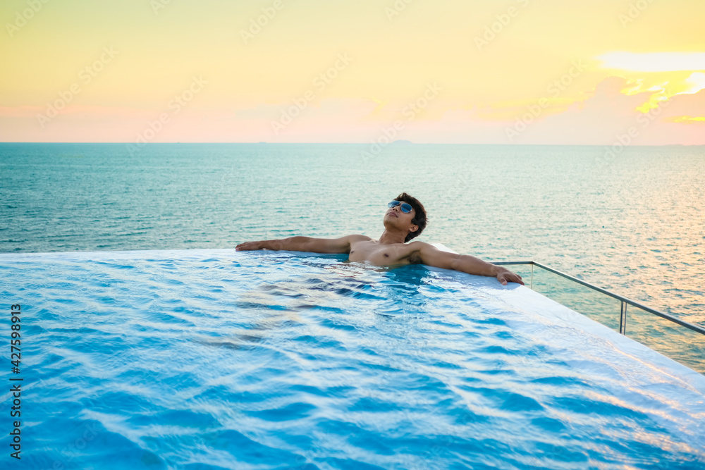 A man relaxing in a swimming pool at a luxury resort with blue sky and blue sea background. Young man enjoying beautiful sunset in luxury hotel near swimming pool. Calmness and serenity. 