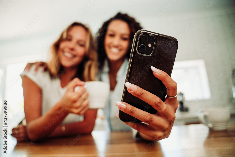Fototapeta premium Beautiful caucasian and mixed race friends sitting in kitchen taking selfie on cellular device drinking hot coffee