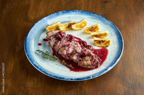 Delicious baked confit duck breast with berry sauce and potatoes, served on a blue plate. Wood background