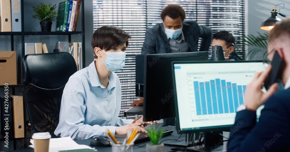 Office workers in quarantine. Rear of male speaking on smartphone while woman worker in medical mask typing on computer in cabinet. Mixed-races males working behind. Profession concept Office workers in quarantine. Rear of male speaking on smartphone while woman worker in medical mask typing on computer in cabinet. Mixed-races males working behind. Profession concept
