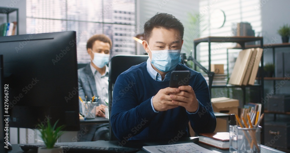 Portrait of happy Asian male employee in medical mask sitting at desk in cabinet texting on smartphone and browsing on computer at workplace in lockdown. Man works in company office, job concept Portrait of happy Asian male employee in medical mask sitting at desk in cabinet texting on smartphone and browsing on computer at workplace in lockdown. Man works in company office, job concept