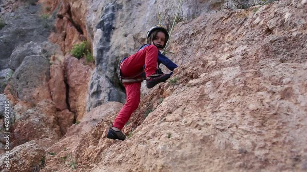 The child is climbing on a natural terrain. A boy in helmet climbs a rock on a background of mountains. Extreme hobby. Athletic kid trains to be strong. Rock climbing safety. The child is climbing on a natural terrain. A boy in helmet climbs a rock on a background of mountains. Extreme hobby. Athletic kid trains to be strong. Rock climbing safety.