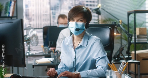Fototapeta Naklejka Na Ścianę i Meble -  Portrait of young Caucasian beautiful woman in medical mask working typing on computer while speaking in headset sitting at table at office. Call center operator talking in cabinet, customer support