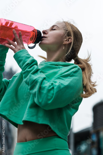 Atractive mature woman wearing activewear is resting after doing exercise and drinking water from a bottle.