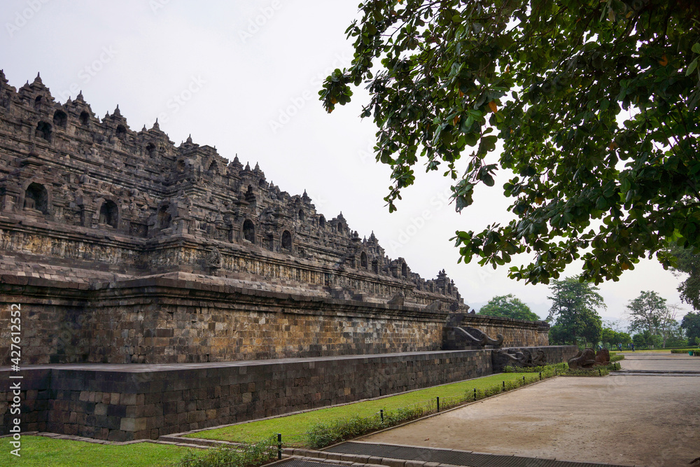 Exterior wall of ancient Borobudur temple view from the base of temple with trees in foreground and background. No people. Popular tourist and Buddhist pilgrimage destination.