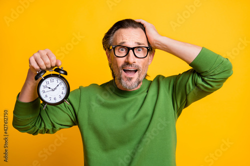 Photo of shocked guy hold alarm clock open mouth hand head wear glasses green sweater isolated yellow color background photo