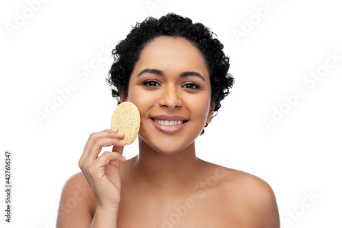 beauty, people and skincare concept - young african american woman with bare shoulders cleaning face with exfoliating sponge over white background