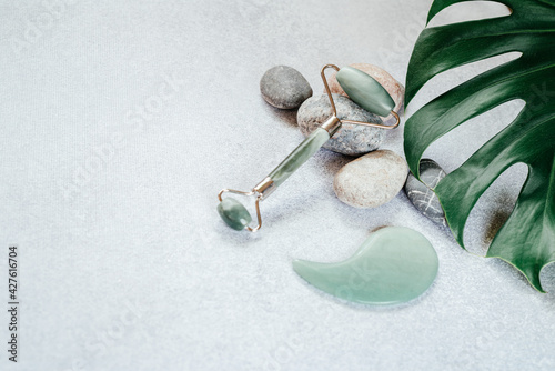Fototapeta Naklejka Na Ścianę i Meble -  Facial massage tools: jade guasha massage roller and scraper with pebble stones and tropical monstera leaf, SPA relax, face and skin care background