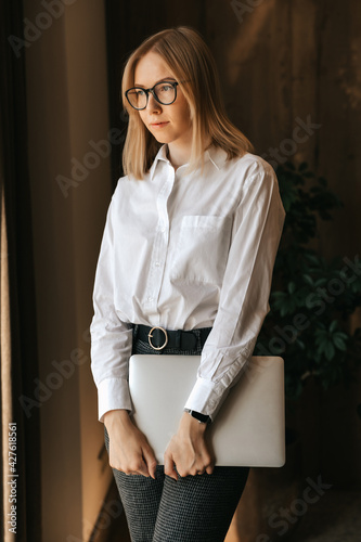 A beautiful business girl in a white shirt and glasses stands with a laptop in her hands in the office against the background of a blackboard with a graph of the company's growth
