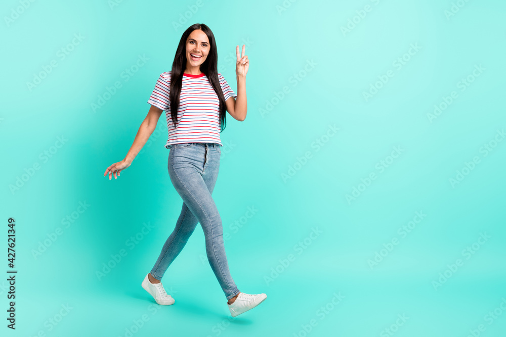 Full size photo of nice brunette optimistic long hairdo lady go show v-sign wear t-shirt jeans sneakers isolated on teal color background Full size photo of nice brunette optimistic long hairdo lady go show v-sign wear t-shirt jeans sneakers isolated on teal color background