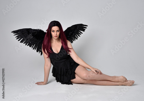 Full length portrait of a red haired girl wearing black dress and feather angel wings. Sitting pose against a studio background. 