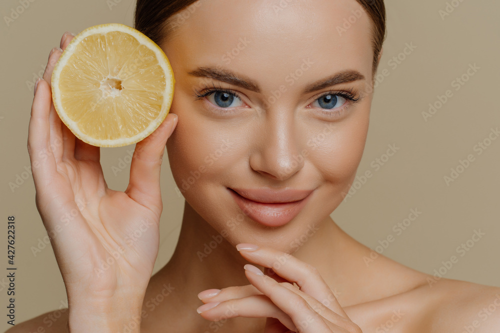 Beauty portrait of healthy young topless brunette woman touches chin gently holds half of juicy fresh lemon has natural makeup poses topless isolated over brown background. Facial care concept Beauty portrait of healthy young topless brunette woman touches chin gently holds half of juicy fresh lemon has natural makeup poses topless isolated over brown background. Facial care concept