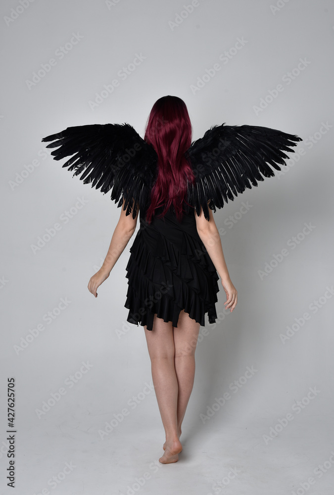 Fototapeta premium Full length portrait of a red haired girl wearing black dress and feather angel wings. Standing pose against a studio background.