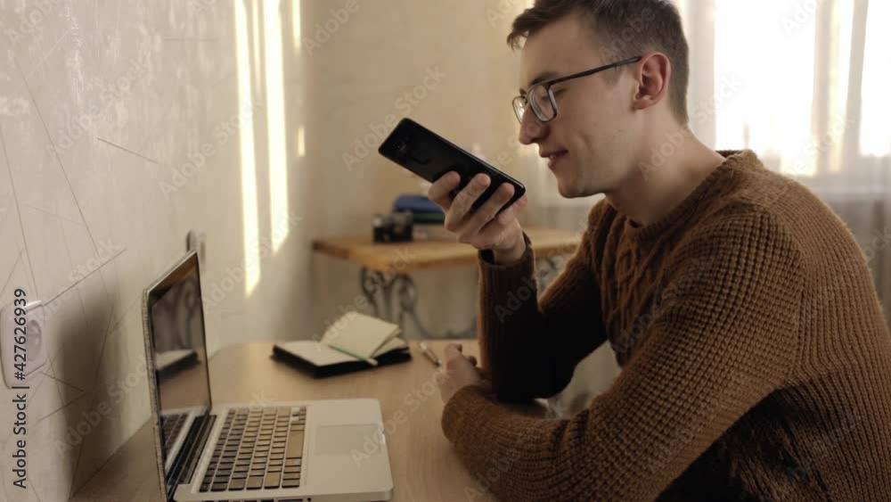 Side view of man recording voice message on smartphone and communicating on social media with friend while sitting at table at home Side view of man recording voice message on smartphone and communicating on social media with friend while sitting at table at home