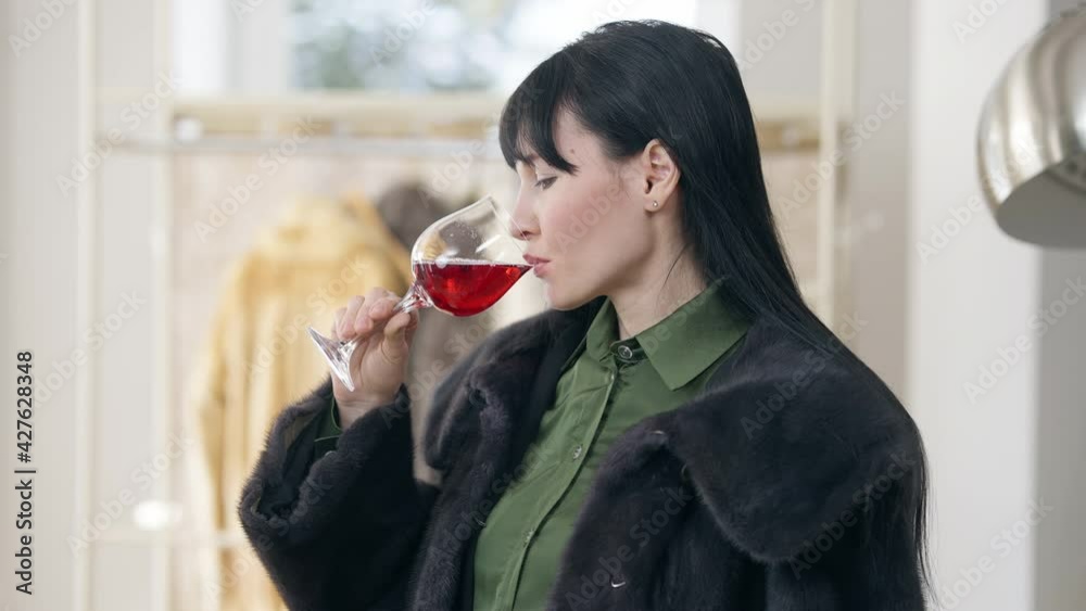Side view of rich young woman in fur coat tasting luxurious red wine indoors. Portrait of serious confident Caucasian wealthy lady drinking beverage thinking. Lifestyle and luxury Side view of rich young woman in fur coat tasting luxurious red wine indoors. Portrait of serious confident Caucasian wealthy lady drinking beverage thinking. Lifestyle and luxury