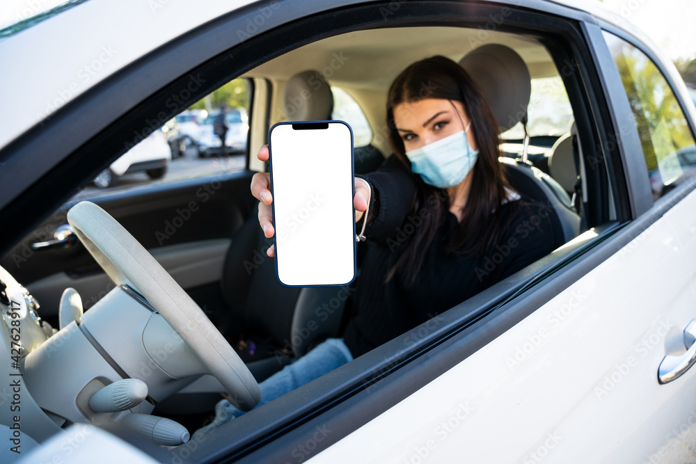 Young woman shows her smartphone at the parking in car on road wearing protective face mask against Coronavirus Covid-19 infection - Mockup of the isolated white blank screen of an electronic device