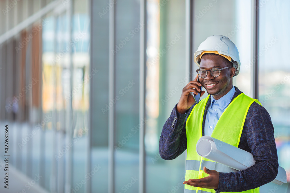 Smiling architect in suit and with helmet on head standing in building in construction process next to window and talking on smart phone. Engineer, architect or supervisor on a construction site Smiling architect in suit and with helmet on head standing in building in construction process next to window and talking on smart phone. Engineer, architect or supervisor on a construction site