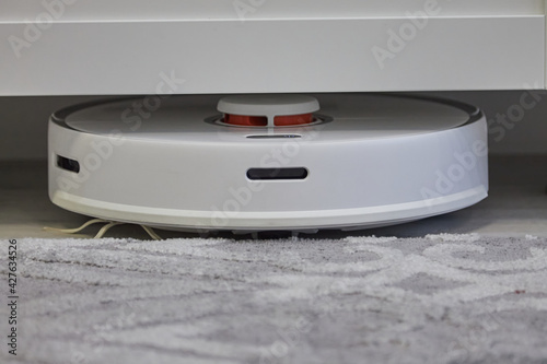 A white robot vacuum cleaner cleans under the cabinet. Smart Home Technology